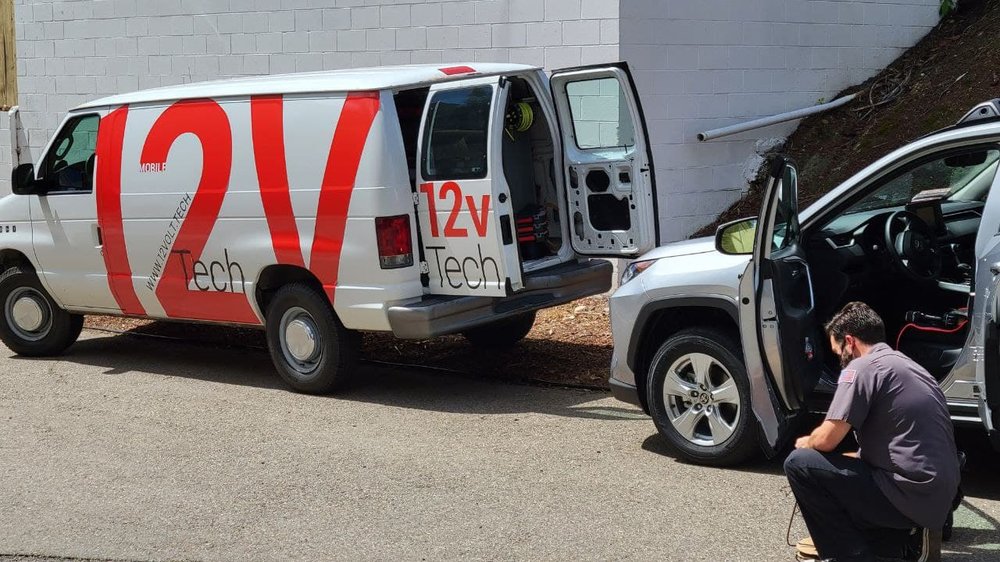 Van with '12V Tech' branding next to a car with open door, on a street.